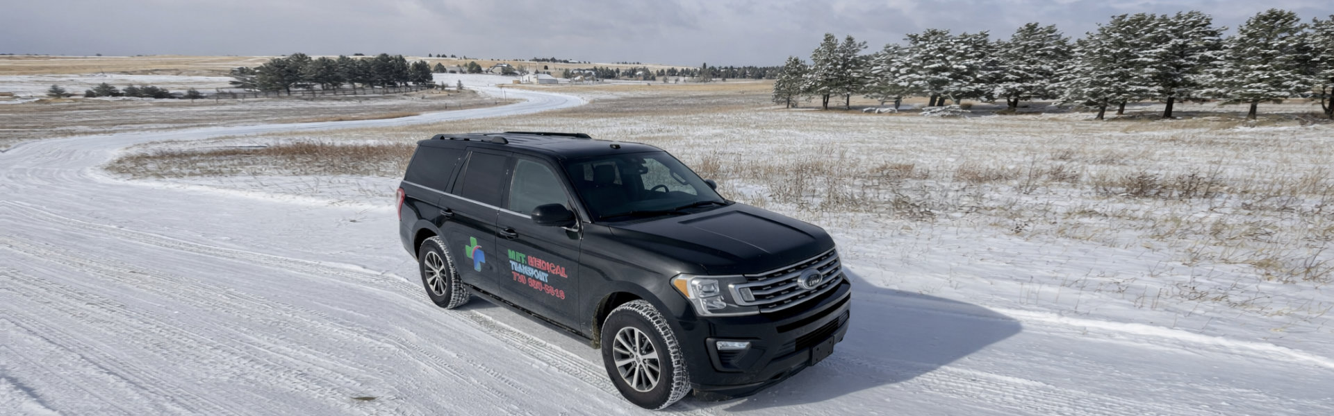 A black SUV drives on a snowy road through a winter landscape with trees and fields